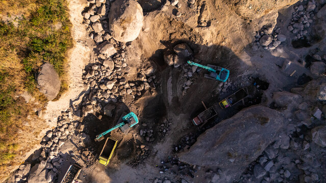 Aerial view of excavators and trucks working in a stone quarry with large boulders and rocky terrain in Klaten, Central Java, Indonesia.