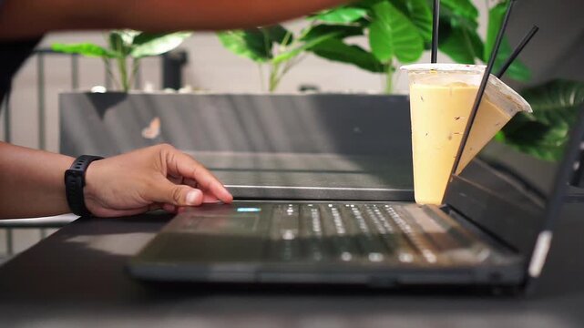 Close-up of hand using laptop on desk in modern cafe with iced coffee and green plants on background, remote work concept
