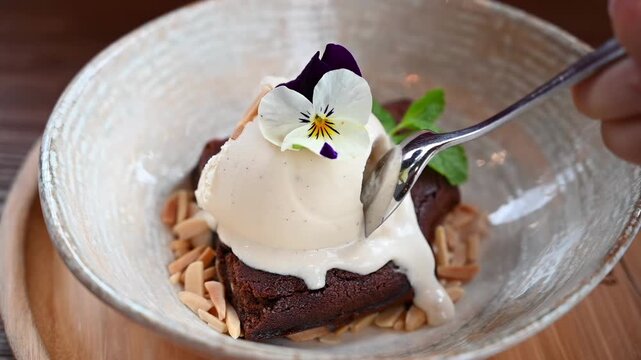 Spoon taking bite of chocolate brownie with vanilla ice cream and edible flower garnish in slow motion.