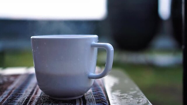 Steaming hot cup of morning coffee or tea in a white mug on an outdoor table during rainy chilly day