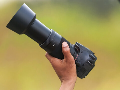 Hand holding professional DSLR camera with long telephoto lens and lens hood isolated on blurred natural background