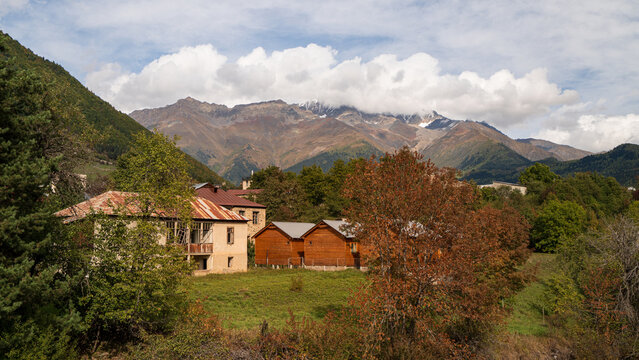 Traditional stone and wooden houses set among autumn trees below snow-capped Caucasus peaks near Mestia, Svaneti, Georgia.