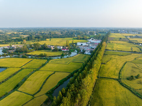 Aerial perspective shows patchwork farmland, a village, tree lines and waterways in a rural landscape under clear sky.