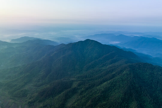 Aerial view of layered forested mountain ranges shrouded in soft mist at dawn, with distant hazy peaks under a pale sky.