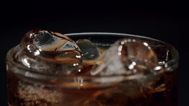 Close-up view of ice cubes slowly swirling and melting in a glass of fizzy cola against a dark background. The refreshing, bubbly soda moves around the cold, clear ice in a dynamic display.