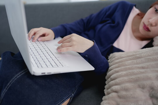 Young Asian woman relaxing on sofa using laptop for remote work from home in casual attire