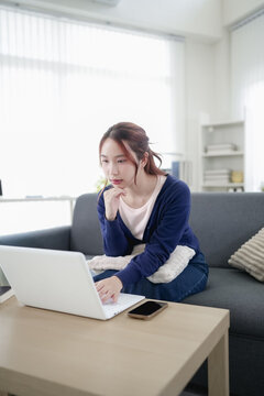 Young Asian woman working on laptop at home sitting on sofa in bright modern living room with copy space