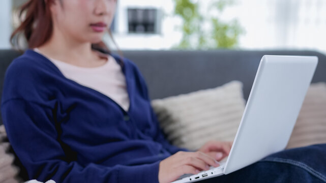 Young Asian woman working on laptop from cozy home sofa in casual blue sweater during hybrid workday