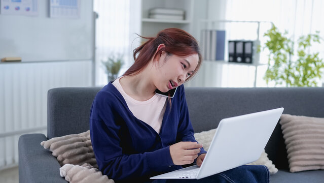 Young Asian businesswoman multitasking on phone call while using laptop on living room sofa at home