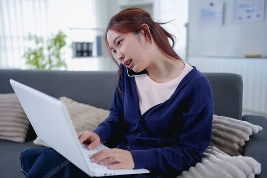 Young Asian woman multitasking on phone call while typing on laptop working from home sofa