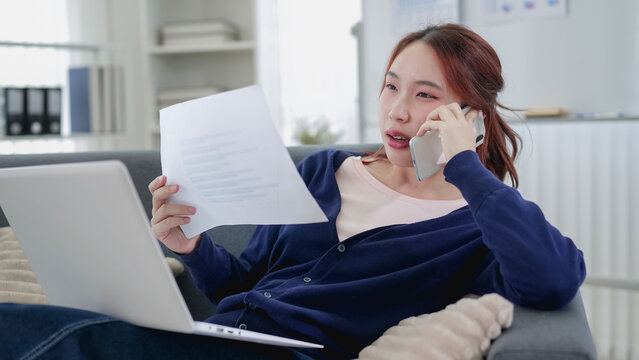 Young Asian businesswoman discussing document over phone call while working from home with laptop on couch