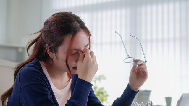Exhausted young Asian woman touching nose bridge experiencing headache stress while working from home office with copy space