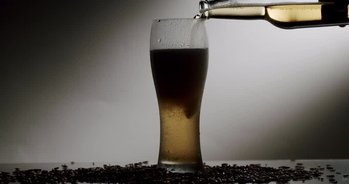 Slow motion pouring of golden beer from a bottle into a cold, misty glass, with malt grains scattered around its base, creating a swirling foam head under a dramatic light