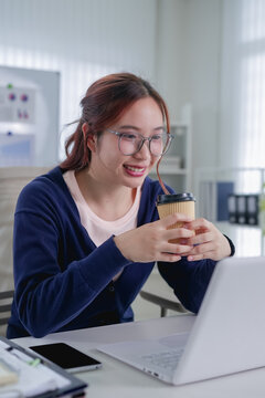 Young Asian businesswoman enjoying coffee during hybrid work break in bright modern office with laptop