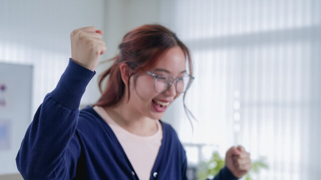 Young Asian businesswoman celebrating success with raised fists while working remotely in bright modern home office