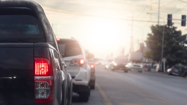 Rear side of pickup car stopped with turn on brake light. Traffic queue on an urban highway under bright sunlight during rush hour. Asphalt road at a crossroads in a rural area with small houses.