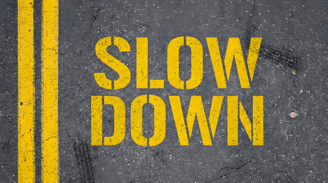 Yellow stencil text reading SLOW DOWN painted on weathered dark asphalt road surface with a double yellow line and small debris scattered around featuring pavement, marking, warning and traffic