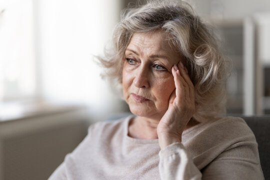 Closeup pensive senior woman sit on couch deep in contemplation, feeling disappointed, touching temple experiencing headache or pressure at home. Mental health issues, stress, or solitude concepts
