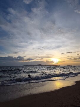 A long vertical shot of a beach sunset with dramatic clouds, a dark sandy shore, small rolling waves, and the silhouette of a lone swimmer in the water near the foreground.