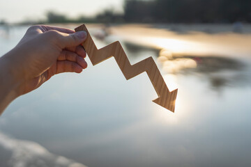Hand holding wooden downward arrow graph at sunset beach background representing financial crisis,...