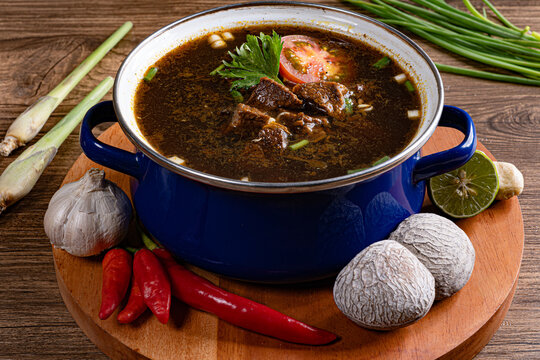 Spicy rawon beef soup plated on round wooden board surrounded by fresh chili and lemongrass