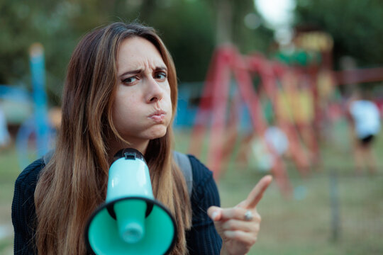 Stressed Mother With Puffed Cheeks Holding Megaphone At Children Playground. Annoyed young mom shows frustration while using a bullhorn outdoors in park