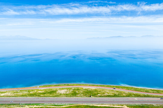 Aerial view of Sayram Lake beside a highway in Xinjiang, China, with blue water, distant mountains, green shoreline, and clear daytime sky.