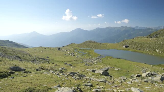 Pan: Alpine landscape in Val Varaita, Piedmont, Italy, featuring a small mountain lake surrounded by rocky grasslands, with layered peaks beneath a clear blue summer sky
