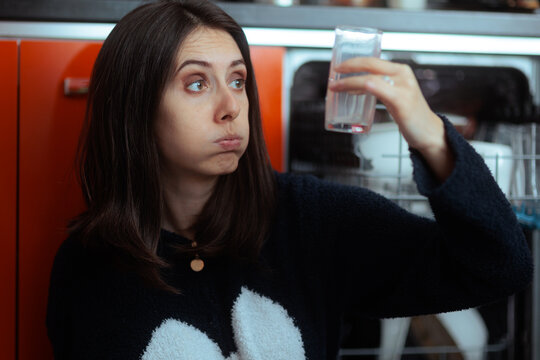Disappointed Young Woman Inspecting Dirty Glass After Poor Dishwasher Clean. Unhappy housewife looks closely at a dirty glass from her bad dishwasher
