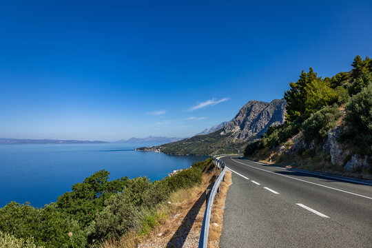 An asphalt road winds along steep cliffs above the Adriatic Sea in Croatia on a sunny summer day. The scene features rocky terrain, Mediterranean vegetation, and a clear blue sky.