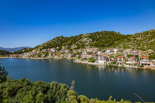 A hillside village with Mediterranean-style houses overlooking a calm bay on the Adriatic coast. The landscape features rocky terrain, green vegetation, and clear blue sky.