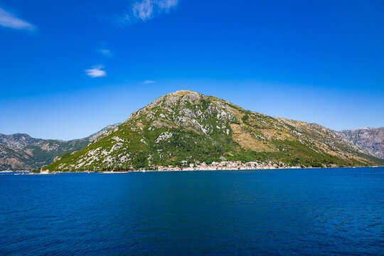 A coastal settlement sits below a large green mountain alongside the blue sea in Kotor Bay under a clear sky.