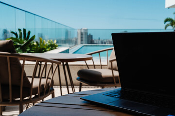 Fototapeta premium Laptop on a terrace overlooking a rooftop pool and city skyline. Workation combines remote work with travel for productivity, flexibility and lifestyle balance