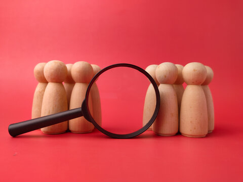 A black magnifying glass rests between two distinct groups of wooden peg figures on a red background