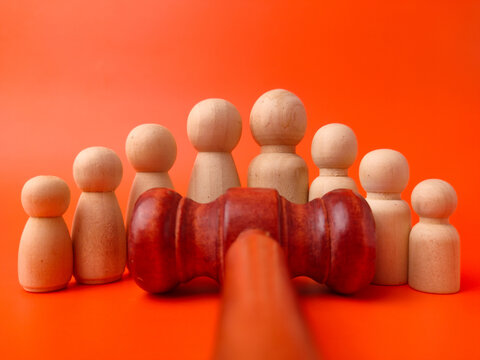 A wooden gavel lies positioned horizontally in front of seven standing wooden peg figures on an orange background
