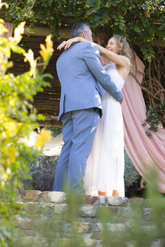 Wedding couple wearing gray suit and lace-dress embracing on low wall under pergola with pink-drape