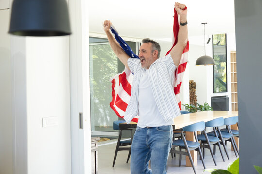 Middle-aged man holding American flag overhead, stepping into modern dining area near wooden table