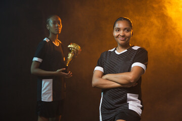 Fototapeta premium African American female team posing in studio, black-white jerseys and shorts, holding gold trophy