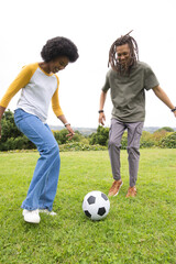 Naklejka premium African American friends kicking black-white soccer ball on grassy slope showing bracelets