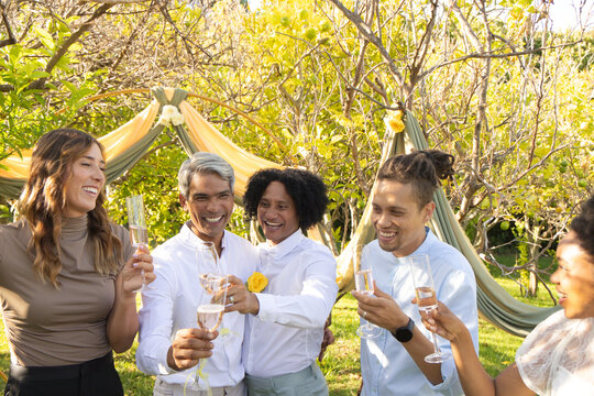 Diverse friends in light outfits toasting champagne flutes on lawn under fabric arch yellow rose