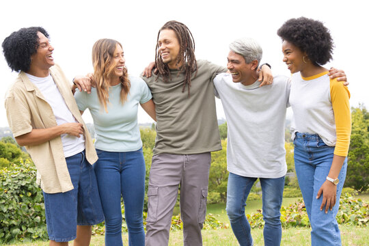 Diverse friends linking arms on grassy hill, wearing T-shirts and jeans with wristwatch visible