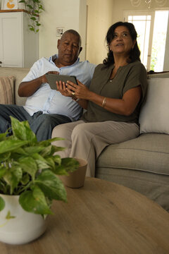 African American senior couple sitting on light sofa at home, holding tablet, wearing blue shirt