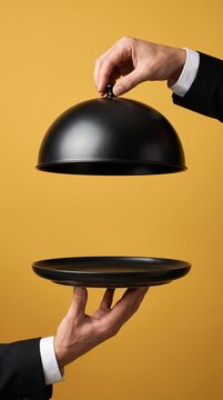 Hands lifting black serving cloche dome cover over empty platter on yellow background