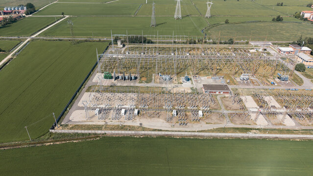 Aerial view of the C.P. Lacchiarella electrical substation featuring high-voltage transformers, switchgear, and power lines surrounded by lush green agricultural fields in Badile, Lombardia, Italy.