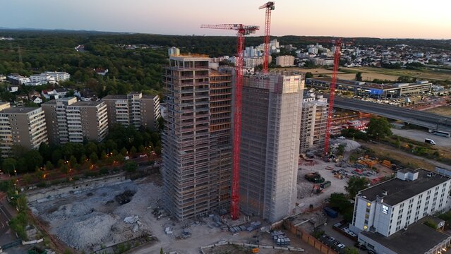 Aerial view of construction site with two tall unfinished high-rise buildings and red tower cranes surrounded by residential blocks and a highway in Offenbach am Main, Hessen, Germany.