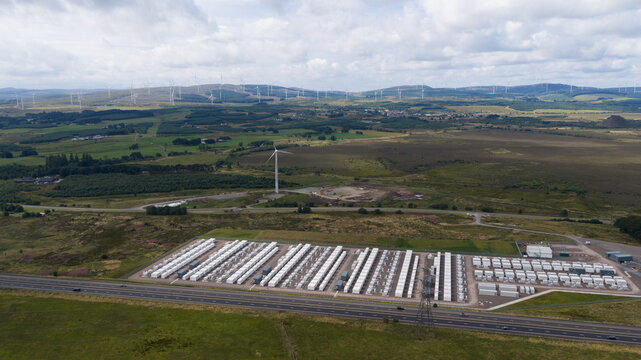 Aerial view of BESS battery energy storage system with rows of white containers and a nearby wind turbine in a rural landscape near Lanark, Scotland, United Kingdom.