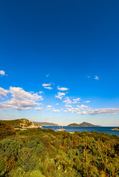 Lush green mediterranean vegetation covers hills overlooking a calm blue sea with islands and a sailboat under a clear sky