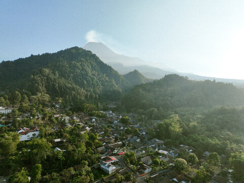 Aerial view of Kaliurang village at the foot of Mount Merapi volcano, featuring lush tropical forests, traditional rooftops, and misty slopes in Kecamatan Pakem, Daerah Istimewa Yogya, Indonesia.
