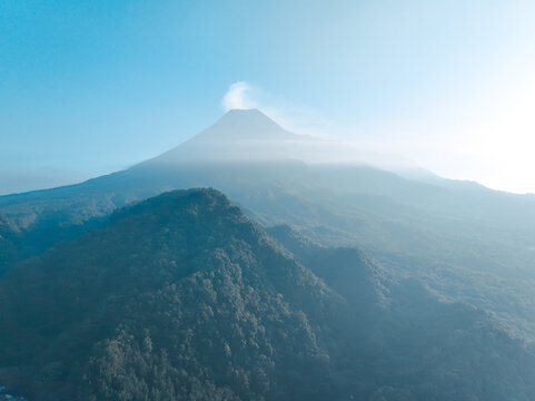 Aerial view of the majestic Mount Merapi volcano rising above lush forested hills with a light mist and clear blue sky in Kecamatan Pakem, Daerah Istimewa Yogya, Indonesia.