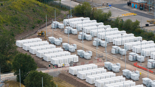 Aerial view of a large-scale battery energy storage system with rows of white containers on a gravel site in Warrington, England, United Kingdom.